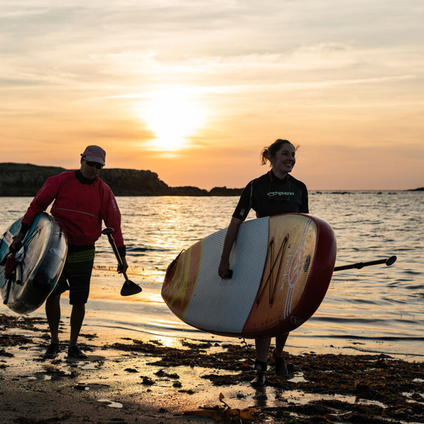 Un homme et une femme avec une planche de paddle sous le bras sur la plage au coucher de soleil